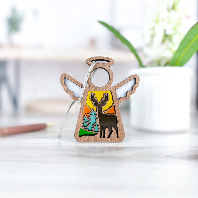 The Autumn Deer Ornament by Forged Flare® is a 3.5" Mother's Angels® wooden angel figurine featuring a deer and pine trees cutout, set against a yellow background with a ribbon loop—ideal as an autumn accent or deer hunter gift.