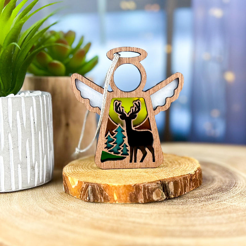 The Forged Flare® Autumn Deer Ornament is a 3.5" Mother's Angels® figurine, featuring an angel with deer, trees, and full moon cutouts, displayed on a wood slice beside a potted plant and blue background.