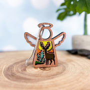 The Autumn Deer Ornament | 3.5" (H) Angel Figurine by Forged Flare® features a cutout deer, pine trees, and a sunset—ideal for autumn decor or as a deer hunter gift. Part of the Mother's Angels® collection. Displayed on wood with blurred plant background.