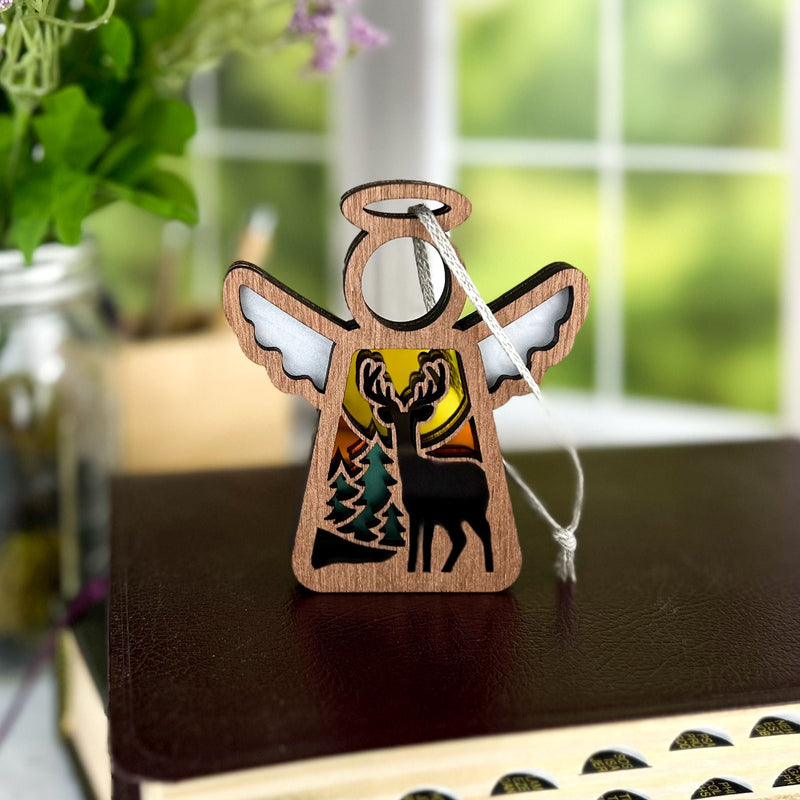The Autumn Deer Ornament by Forged Flare® is a 3.5"H Mother's Angels® figurine with a deer and sunset cutout, resting on a book. Perfect as an autumn or deer hunter gift, it features a silver string and blurred greenery in the background.