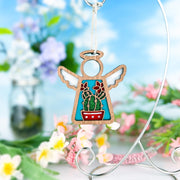 The Forged Flare® Desert Cactus Ornament | 3.5" (H) Mother's Angels® angel figurine is displayed on a silver stand among pink, white, and purple blooms, with a blurred outdoor backdrop of blue sky and clouds.