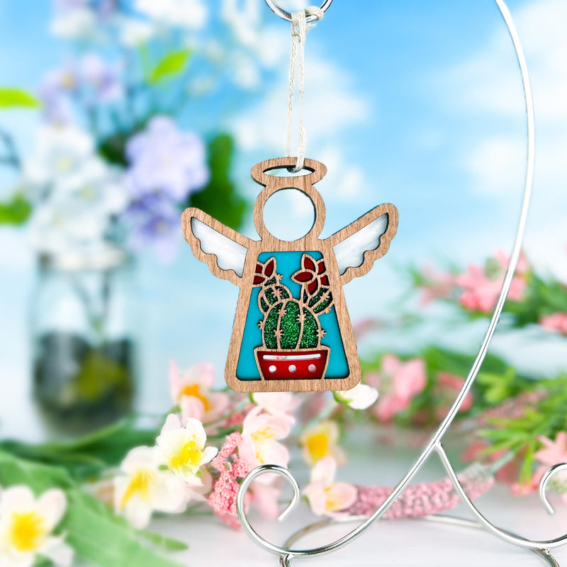 The Forged Flare® Desert Cactus Ornament | 3.5" (H) Mother's Angels® angel figurine is displayed on a silver stand among pink, white, and purple blooms, with a blurred outdoor backdrop of blue sky and clouds.