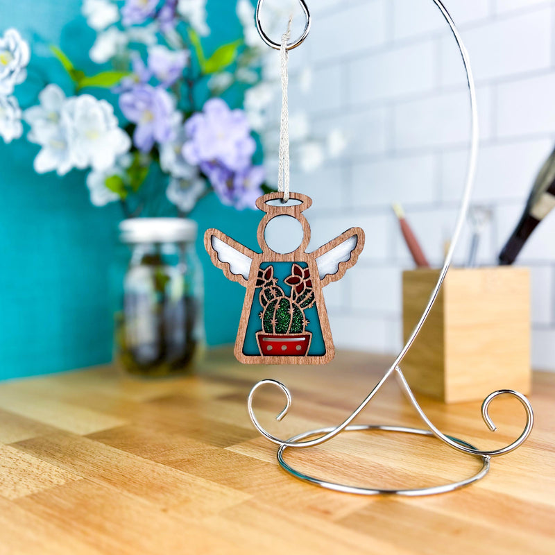 The Desert Cactus Ornament, a 3.5" angel figurine from Mother's Angels® by Forged Flare®, hangs from a metal stand on a wooden counter with flowers, a jar, and pencils seen against a teal and white tiled wall in the background.