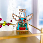 The Forged Flare® Mother's Angels® Desert Cactus Ornament is a 3.5" wooden angel figurine, featuring red cactus flowers in a pot on turquoise, hanging by a ribbon for Southwest holiday decor. Blurred festive accents complete the look.