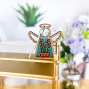 The Forged Flare® Desert Cactus Ornament, a 3.5" Mother's Angels® angel figurine with white wings and a colorful cactus design, sits on a gold shelf. A green succulent and purple-white flowers appear blurred in the background.