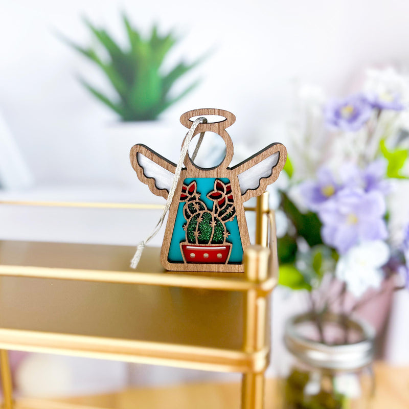 The Forged Flare® Desert Cactus Ornament, a 3.5" Mother's Angels® angel figurine with white wings and a colorful cactus design, sits on a gold shelf. A green succulent and purple-white flowers appear blurred in the background.