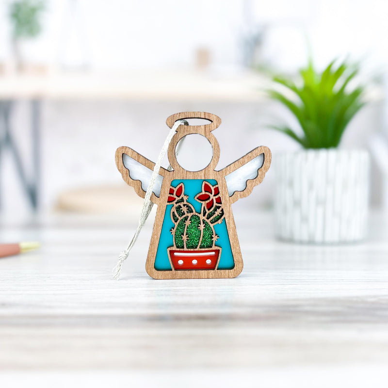 The Forged Flare® Mother's Angels® Desert Cactus Ornament is a 3.5" wooden angel figurine featuring a cactus and red flowers, ideal for Southwest decor. It stands on a light wood surface with a green plant and blurred background behind it.