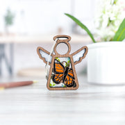 The Forged Flare® Mother's Angels® Monarch Butterfly Ornament with Imperfections (3.5" angel figurine) rests on a light surface with green plants and a white pot behind it—an uplifting, thoughtful memorial gift.