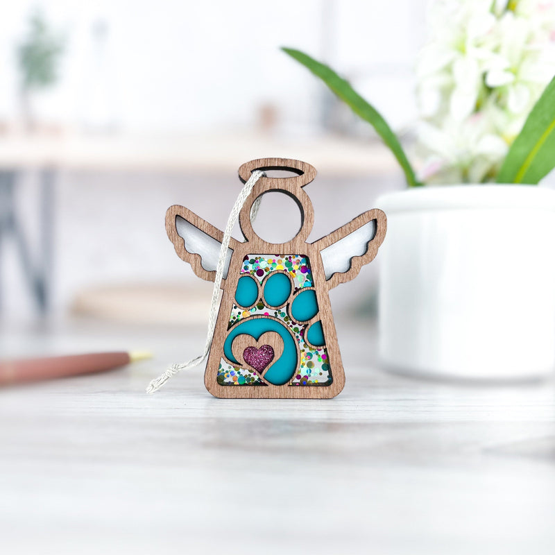The Forged Flare® Paw Print Ornament with Imperfections is a 3.5" wooden Mother's Angels® figurine featuring a heart and paw print—a touching pet memorial gift. It stands on a light surface with blurred greenery in the background.
