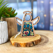 The Forged Flare® Mother's Angels® Paw Print Ornament with Imperfections is a 3.5" wooden angel figurine featuring glittery wings and a heart. It hangs on a white string, making it an ideal pet memorial gift. Potted plant and blurred lights in the background.