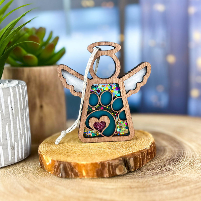 The Forged Flare® Mother's Angels® Paw Print Ornament with Imperfections is a 3.5" wooden angel figurine featuring glittery wings and a heart. It hangs on a white string, making it an ideal pet memorial gift. Potted plant and blurred lights in the background.