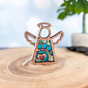 The Forged Flare® Paw Print Ornament with Imperfections is a 3.5" Mother's Angels® angel figurine, featuring turquoise and mosaic details with a glittery pink heart—ideal as a pet memorial gift. Displayed on wood, plant backdrop included.