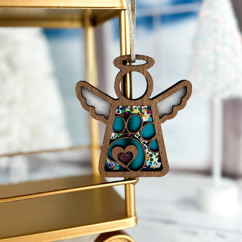 The Forged Flare® Paw Print Ornament with Imperfections, a 3.5" (H) Mother's Angels® angel figurine, hangs from a gold stand amid white trees and snow—an ideal pet memorial gift with colorful mosaic and a blue paw at its center.