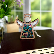 A Forged Flare® Mother's Angels® Paw Print Ornament with Imperfections, a 3.5" wooden angel figurine with white wings and glitter, sits on a closed book. The heart and paw design is ideal for pet memorial gifts. Blurred plants and a window appear behind it.