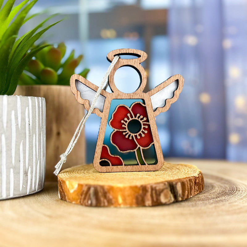The Forged Flare® Mother's Angels® Poppy Ornament (3.5" Angel Figurine) features a red flower design and stands on a tree slice next to a potted plant, with soft blue tones and string lights—ideal for your Amazon marketplace listing.