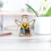 The Forged Flare® Mother's Angels® Winter Deer Ornament, a 3.5" wooden angel figurine, stands on a bright desk near a white cup, green plant, and pen with a painted forest scene featuring a deer, tree, and yellow sky.