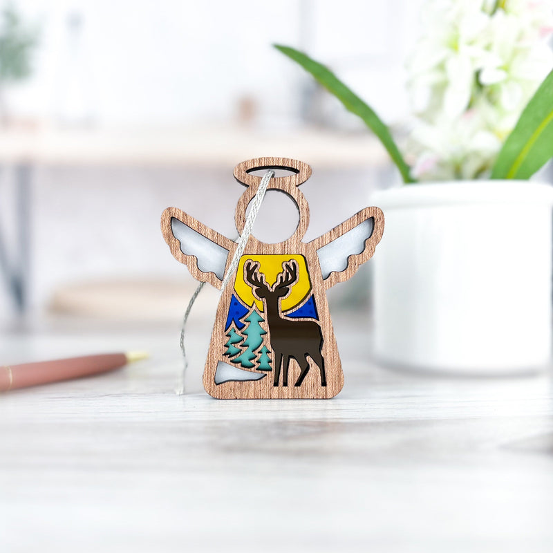 The Forged Flare® Mother's Angels® Winter Deer Ornament, a 3.5" wooden angel figurine, stands on a bright desk near a white cup, green plant, and pen with a painted forest scene featuring a deer, tree, and yellow sky.
