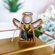 The Forged Flare® Winter Deer Ornament is a 3.5" Mother's Angels® figurine with winged wood design, deer and trees under a moonlit sky, silver hanging string, and gold base—perfect winter decor. Blurred flowers appear in the background.