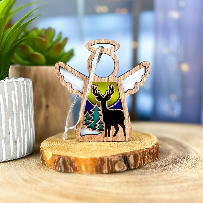The Forged Flare® Winter Deer Ornament | 3.5" (H) Mother's Angels® figurine features a stained glass angel with a winter deer scene, displayed on a tree slice next to a potted plant and blurred background.