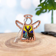 A Forged Flare® Mother's Angels® 3.5" Winter Deer Ornament features a carved wooden angel with wings, a deer cutout, two pine trees, and a yellow sun or moon, set on wood with blurred green leaves in the background.
