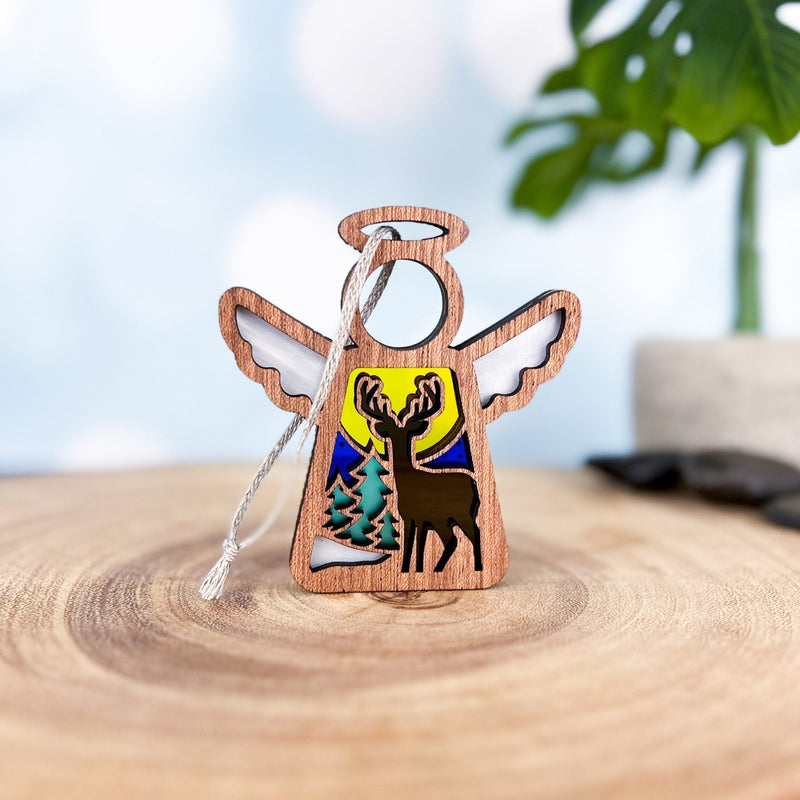A Forged Flare® Mother's Angels® 3.5" Winter Deer Ornament features a carved wooden angel with wings, a deer cutout, two pine trees, and a yellow sun or moon, set on wood with blurred green leaves in the background.