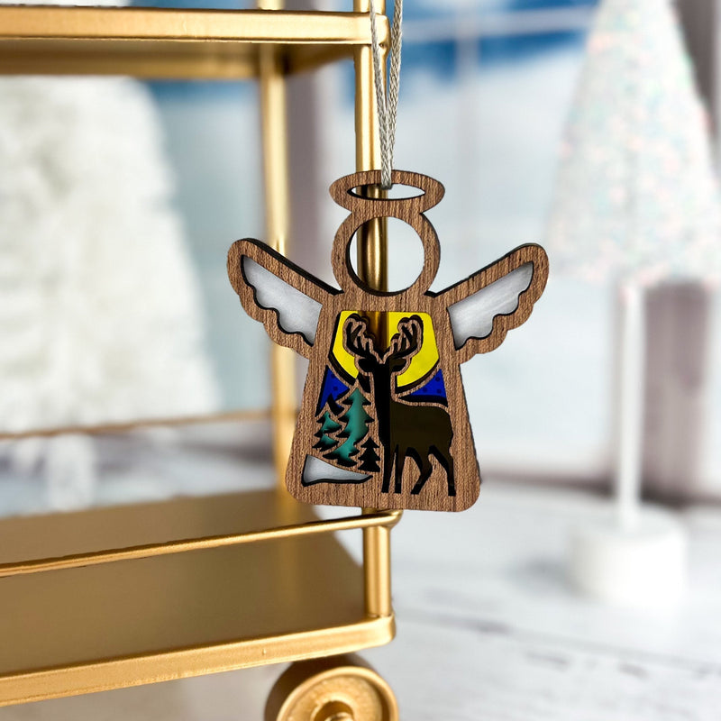 The Winter Deer Ornament | 3.5" (H) Angel Figurine by Forged Flare® from the Mother's Angels® collection hangs before a gold cart, showcasing a deer, trees, and a yellow-blue background amid blurred holiday décor.