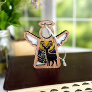 A Forged Flare® Mother's Angels® Winter Deer Ornament, a 3.5" wooden angel figurine with a cutout deer scene and colorful sky, sits atop a closed book with greenery and a window softly blurred in the background.