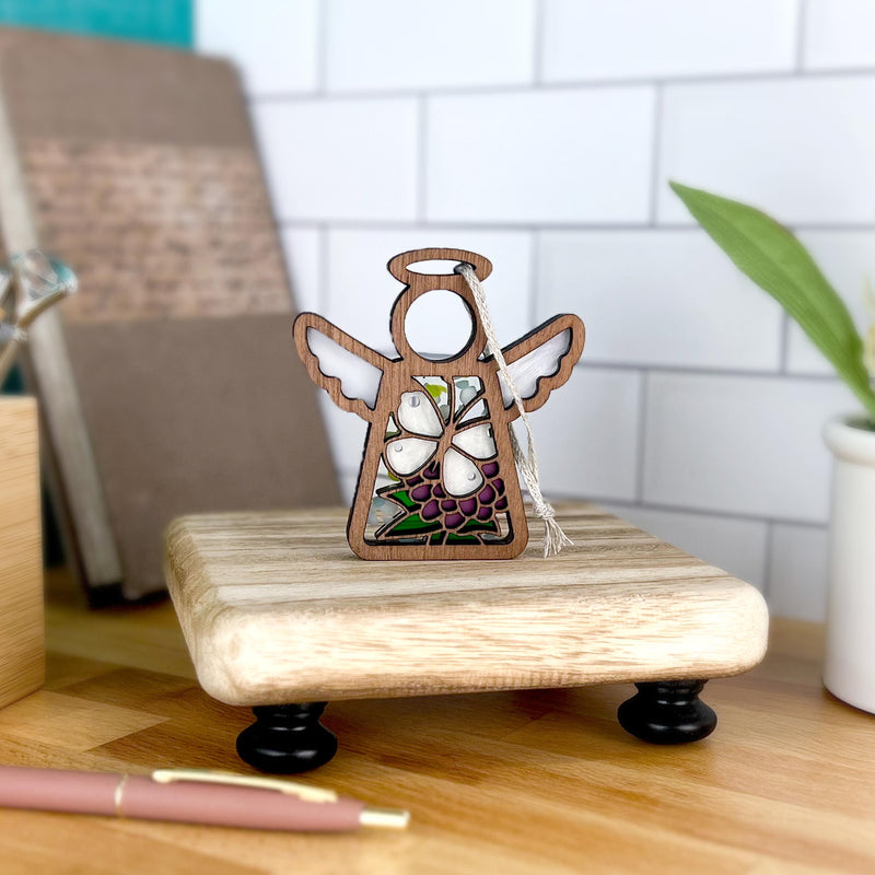 The Forged Flare® Mother's Angels® 3.5" Cabbage Butterfly with Clover Flower Ornament features a wooden angel figurine with stained glass-style wings, shown beside a pen, book, and plant—ideal decor for butterfly and angel lovers.