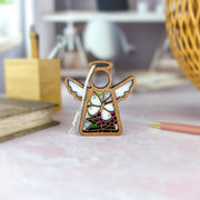 The Forged Flare® Cabbage Butterfly with Clover Flower Ornament, a 3.5" Mother's Angels® figurine, features white wings and stained glass floral accents, displayed on a pink surface with stationery and a softly blurred room in the background.