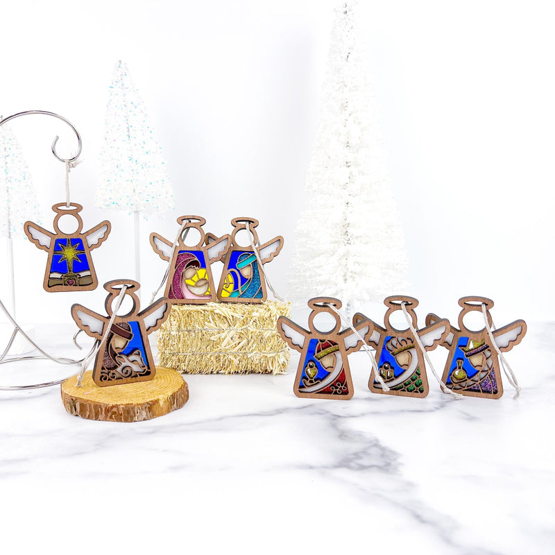 The Forged Flare® Mother's Angels® Christmas Nativity Set Bundle features 3.5" stained glass angel figurines displayed on marble with wood slices, straw, and white Christmas trees in the background.