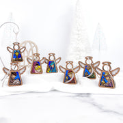 The Forged Flare® Christmas Nativity Set Bundle features seven 3.5" Mother's Angels® figurines with colorful stained-glass designs, displayed on white cloth among festive trees for a serene holiday scene.
