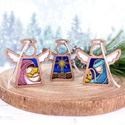 Three Forged Flare® Mother's Angels® figurines from the Christmas Nativity 3-Piece Bundle—Star, Mary, and Joseph—rest on wood. Blurred snowy pines in the background create a festive scene honoring the Holy Family. Each angel stands 3.5" tall.