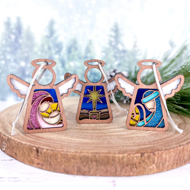 Three Forged Flare® Mother's Angels® figurines from the Christmas Nativity 3-Piece Bundle—Star, Mary, and Joseph—rest on wood. Blurred snowy pines in the background create a festive scene honoring the Holy Family. Each angel stands 3.5" tall.