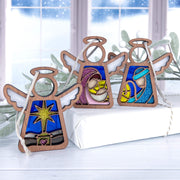 Three Forged Flare® Mother's Angels® 3.5" Nativity angel figurines—Star, Mary, and Joseph—stand atop a white gift box before a snowy window and green foliage for a festive, winter display.