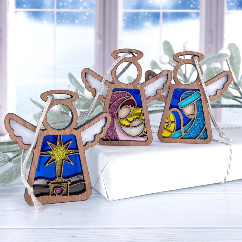 Three Forged Flare® Mother's Angels® 3.5" Nativity angel figurines—Star, Mary, and Joseph—stand atop a white gift box before a snowy window and green foliage for a festive, winter display.