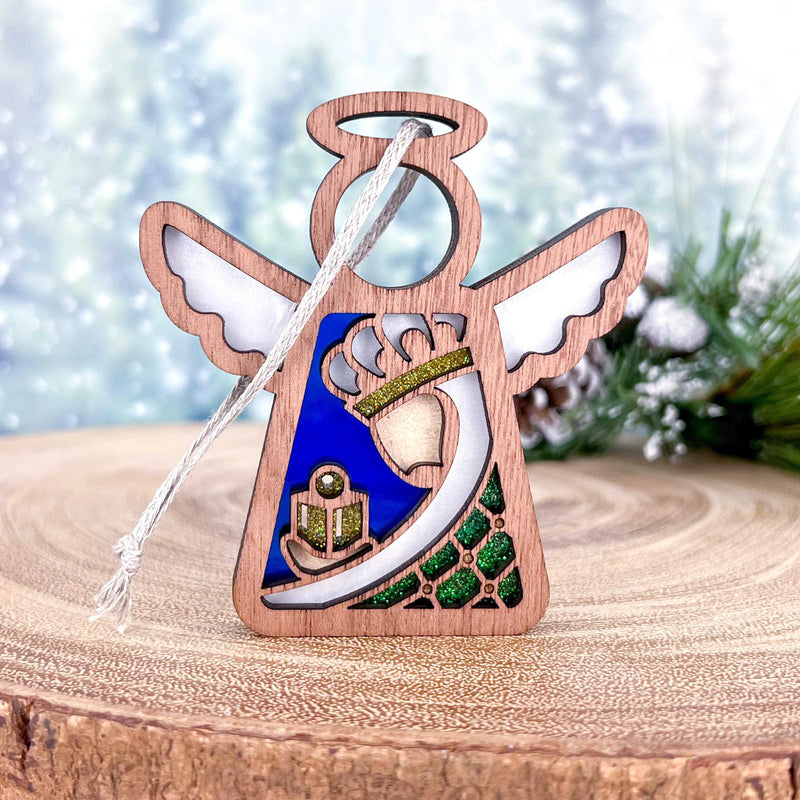 The Christmas Nativity Green Wise Man is a 3.5" angel figurine from Forged Flare® Mother's Angels®, featuring white wings, a blue-green dress with a gold-trimmed bell motif, set on a tree slice with snowy pines and includes a white hanging string.