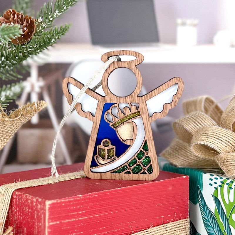 The Forged Flare® Christmas Nativity Green Wise Man 3.5" Angel Figurine hangs on a red gift box, with a Christmas tree branch, burlap bow, and blurred indoor setting in the background.