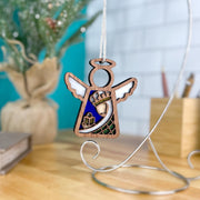 The Christmas Nativity Green Wise Man 3.5" Angel Figurine by Forged Flare® features painted details and hangs on a silver stand, with a blurred plant and books in the background on a wooden surface.