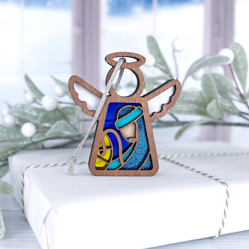 The Forged Flare® Christmas Nativity Joseph 3.5" Angel Figurine from Mother's Angels® features a stained-glass-style design and is displayed on a white box with frosted greenery and a snowy window in the background.