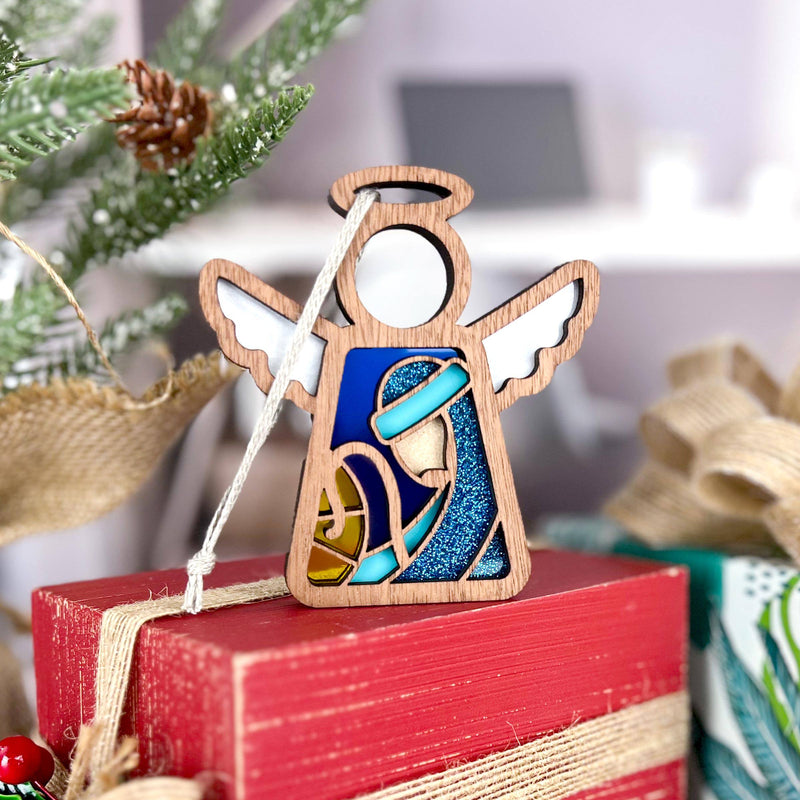 The Forged Flare® Christmas Nativity Joseph Angel Figurine (3.5" H, Mother's Angels®) features blue and gold stained glass-style details, displayed on a red box beside a tree branch—perfect as a festive Nativity gift.