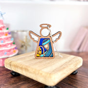 The Forged Flare® Mother's Angels® 3.5" Christmas Nativity Joseph angel figurine stands on a platform, with a softly blurred pink Christmas tree in the background.