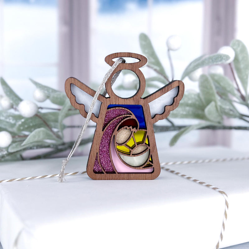 The Forged Flare® Mother's Angels® 3.5" Christmas Nativity Mary Mother of Jesus angel figurine sits atop a white gift box, creating a charming festive gift with holiday greenery and a snowy window blurred in the background.