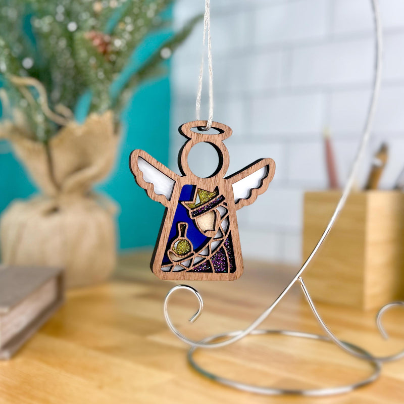 The Christmas Nativity Purple Wise Man 3.5" Angel Figurine by Forged Flare® hangs from a string on a metal stand—ideal as a Nativity gift or for your Christmas Nativity ornament collection. Blurred books and a plant rest in the background.