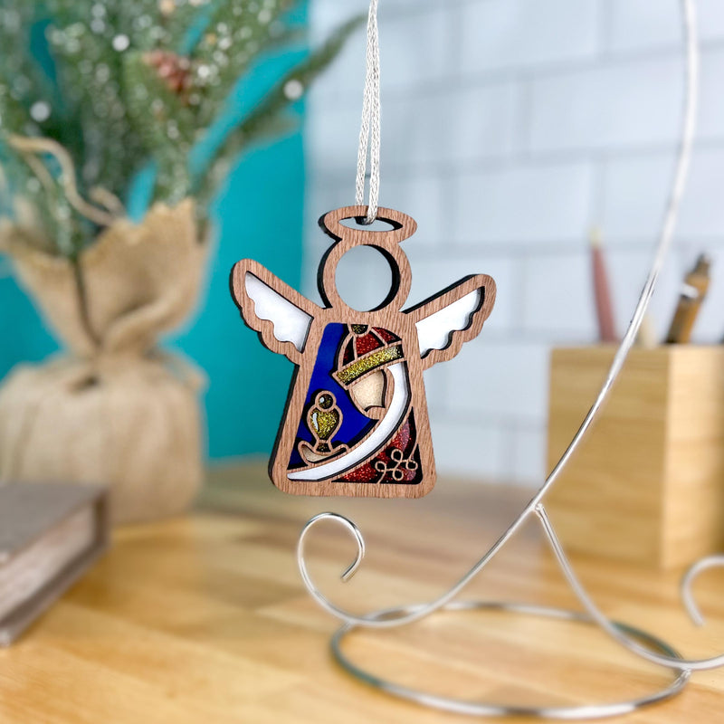 The Forged Flare® Christmas Nativity Red Wise Man Mother's Angels® 3.5" angel figurine, with a crown and gold details, hangs on a silver stand with a blurred plant and tiled wall in the background—a perfect Nativity gift.