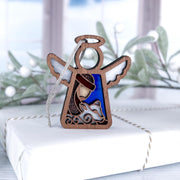 A Forged Flare® Christmas Nativity Shepherd With Sheep 3.5" Angel Figurine from Mother's Angels® sits on a white wrapped Nativity gift, with blurred greenery and a window in the background.