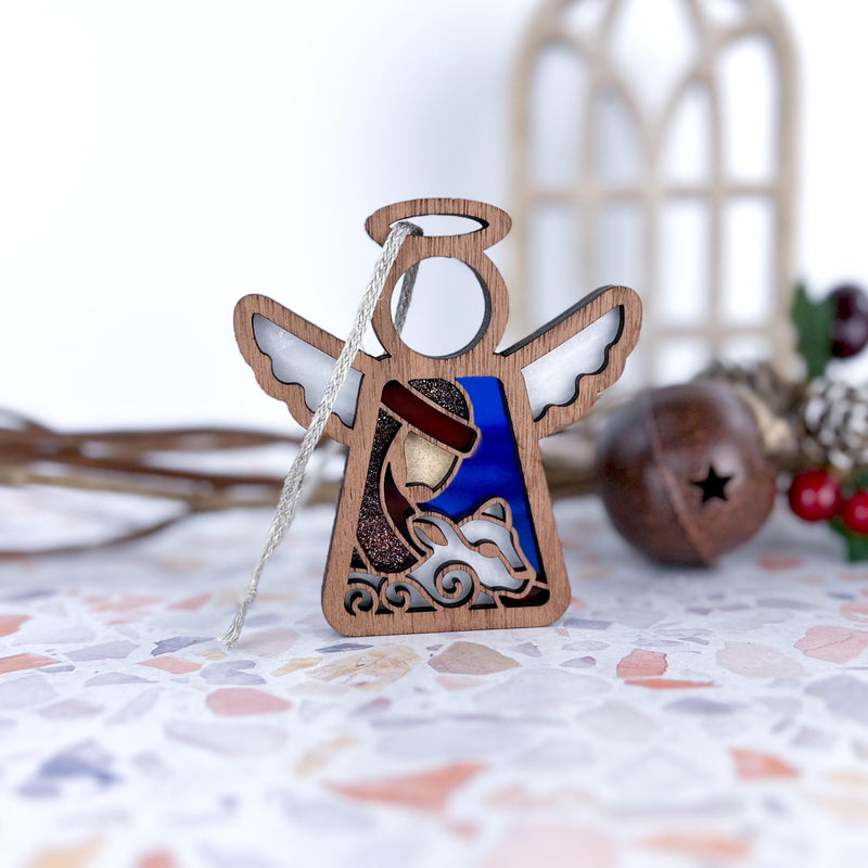 The Forged Flare® Christmas Nativity Shepherd With Sheep 3.5" (H) Angel Figurine from Mother's Angels® features a shepherd and sheep design, white wings, a halo, and hanging string. Holiday décor is softly blurred in the background.