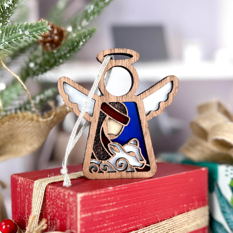 The Forged Flare® Mother's Angels® 3.5" Nativity Shepherd With Sheep figurine sits on a red gift box, making a charming holiday gift. A decorated Christmas tree and wrapped presents add a festive touch to the scene.