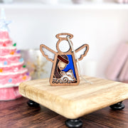 The Forged Flare® Mother's Angels® Christmas Nativity Shepherd With Sheep 3.5" angel figurine is displayed on a small riser before a pink Christmas tree, perfect as a Nativity ornament or holiday gift.