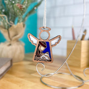 The Forged Flare® Christmas Nativity Shepherd With Sheep 3.5" (H) Angel Figurine from the Mother's Angels® collection hangs from a silver stand on a wooden table, making a charming Nativity gift. Blurred pencils, notebook, and plant are in the background.