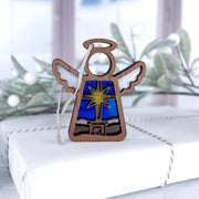 The Forged Flare® Christmas Nativity Star with Imperfections 3.5" Mother's Angels® wooden angel figurine sits on a twine-tied gift box, surrounded by softly blurred holiday décor and frosted greenery for a heartfelt holiday scene.